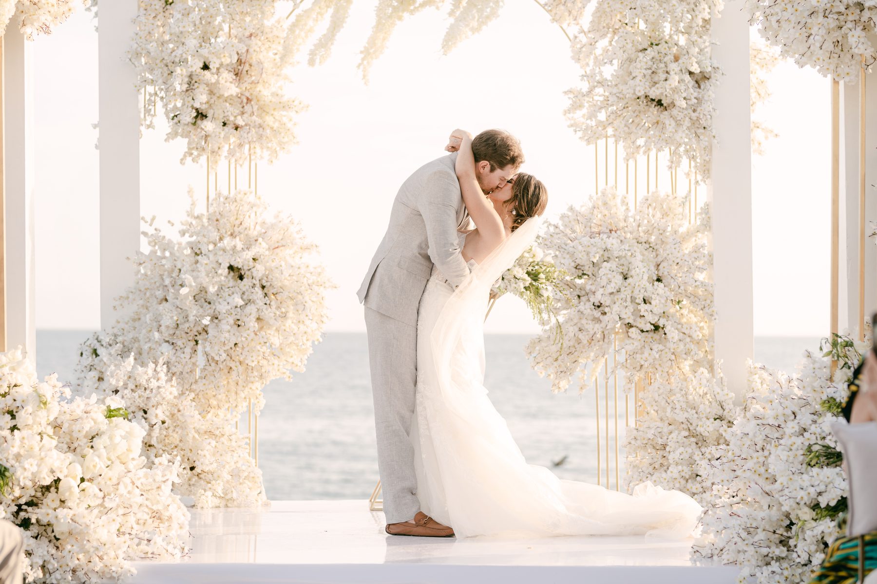 Los novios se besan bajo un arco de flores blancas, frente al mar