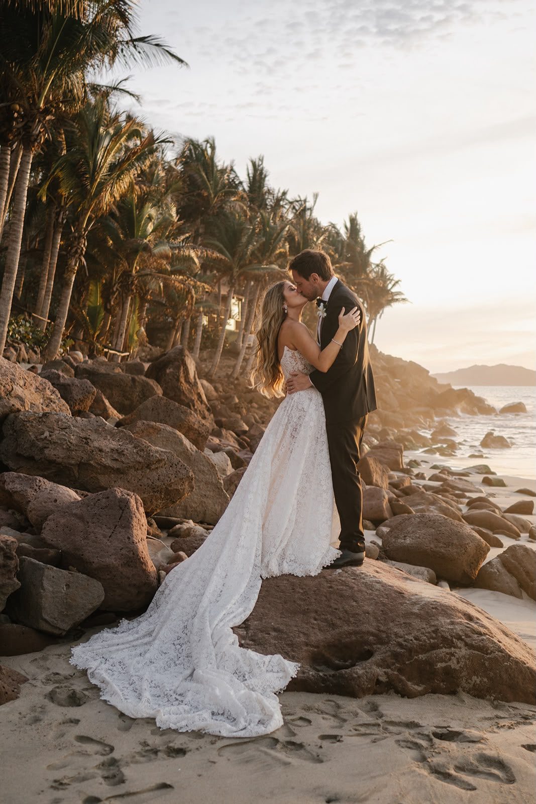 Los novios se besan sobre las rocas al atardecer, entre palmeras